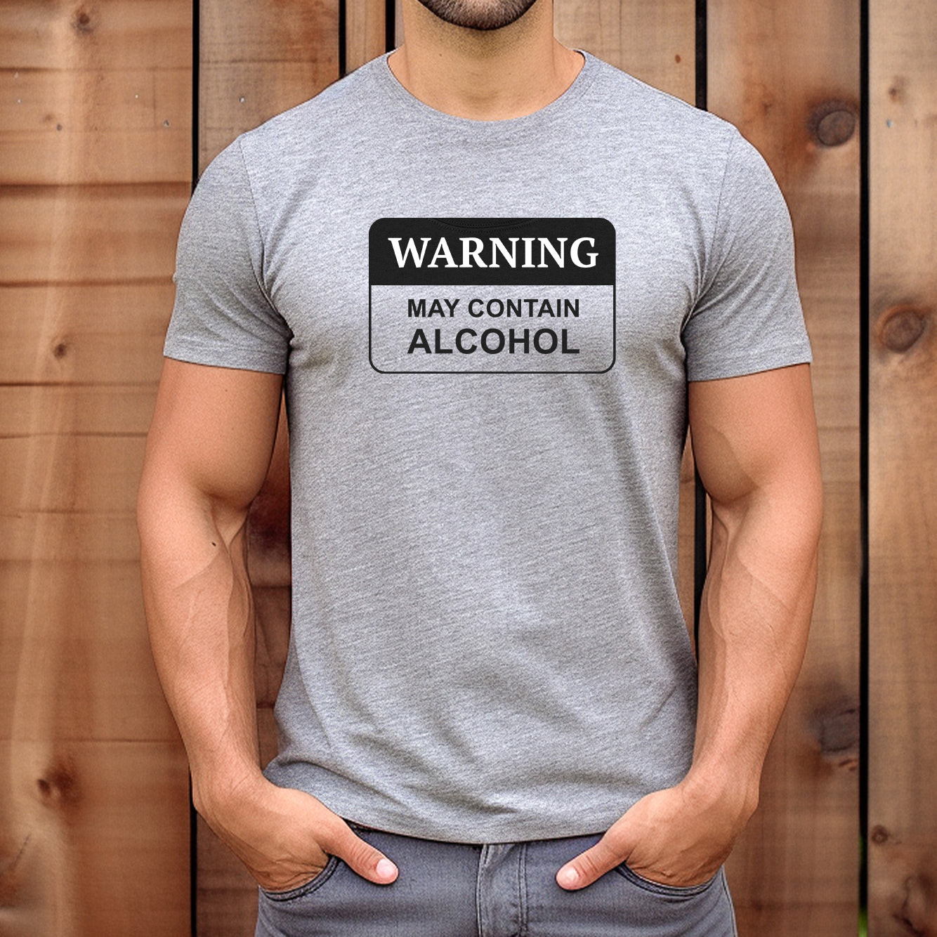 Muscular man standing against a wooden wall with hands in pockets wearing the grey WARNING may contain alcohol graphic tee