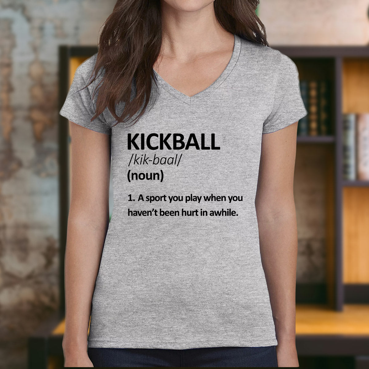 Woman wearing grey v-neck kickball definition shirt with humorous text about playing sports.