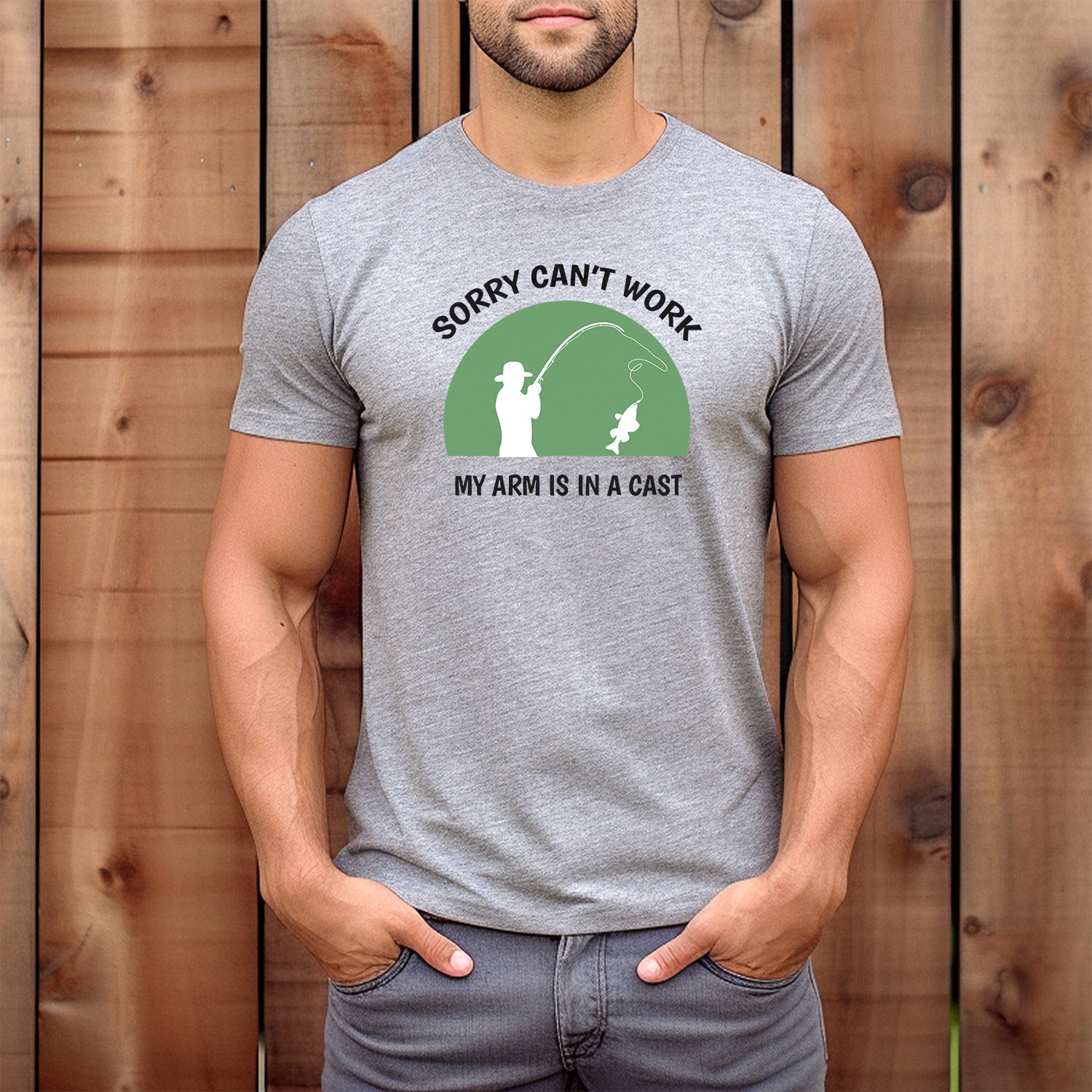 Man wearing heather gray fishing joke T-shirt with green badge, fisherman silhouette, and text Sorry can’t work, my arm is in a cast.