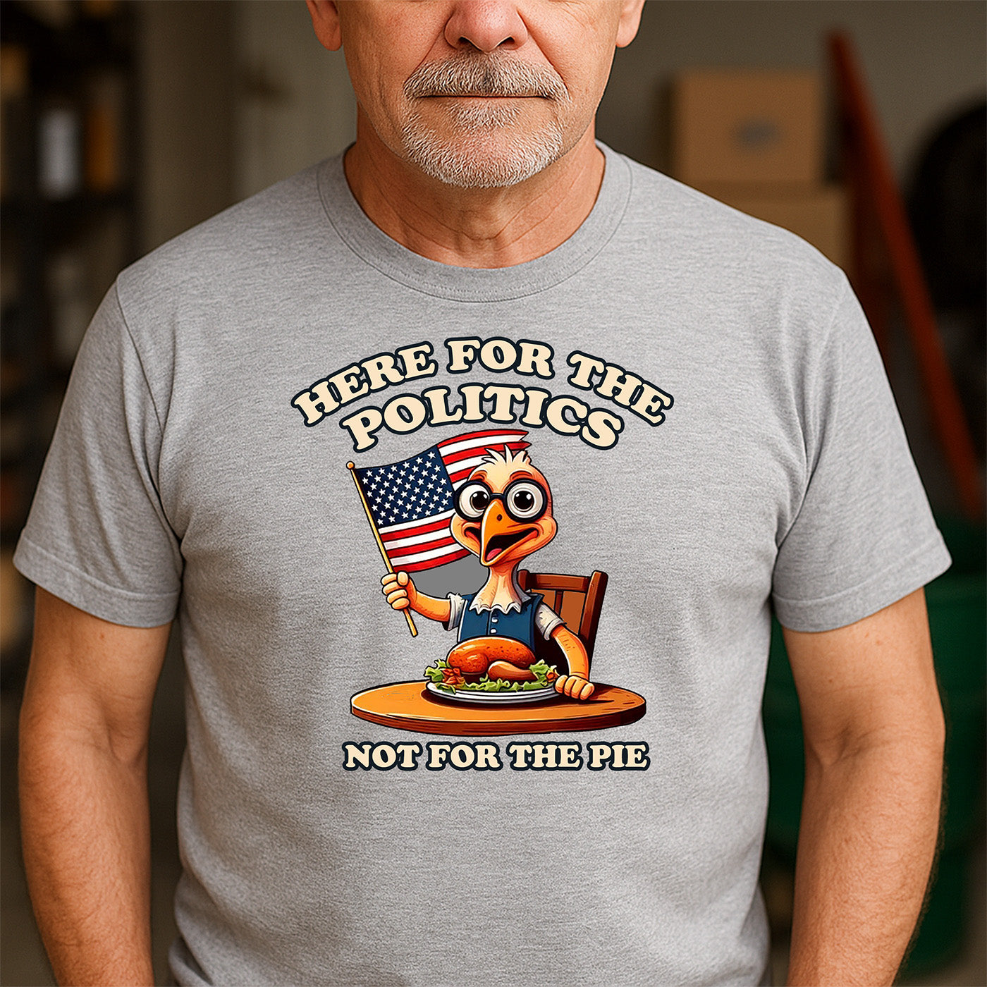 older man wearing gray t-shirt featuring cartoon turkey waving american flag and text here for the politics not for the pie