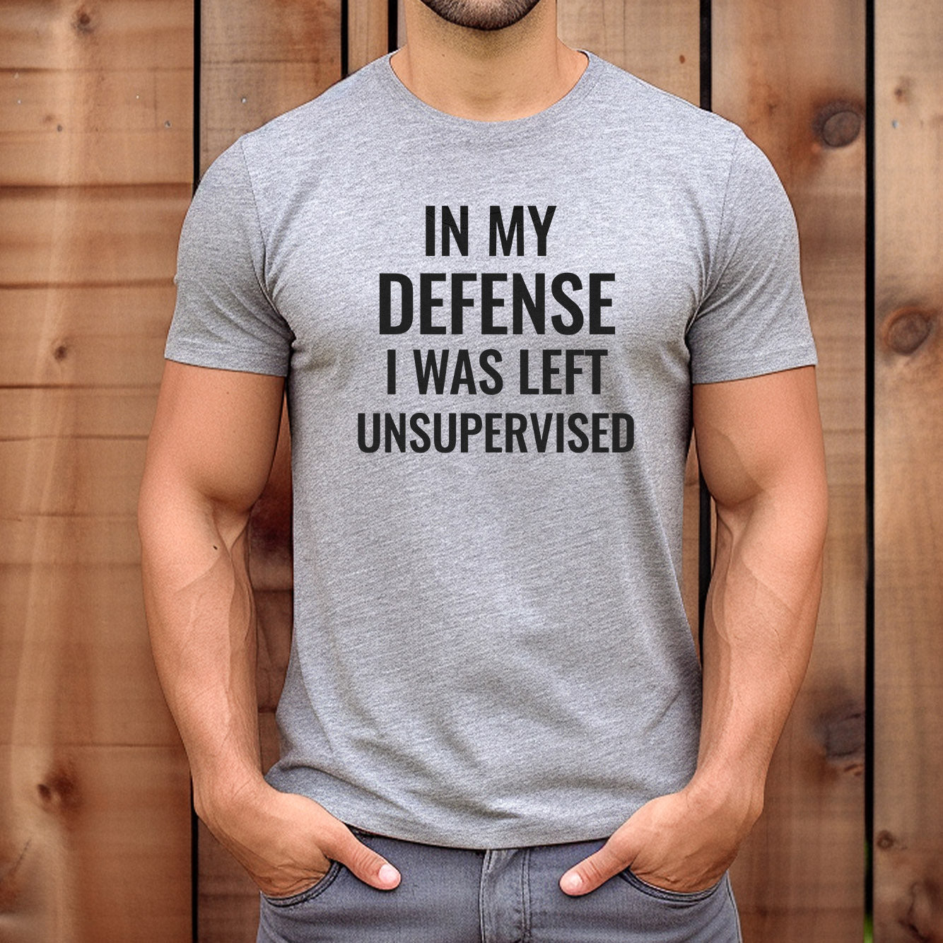 Man wearing grey graphic t-shirt with black text that reads “In My Defense I Was Left Unsupervised,” styled casually outdoors.