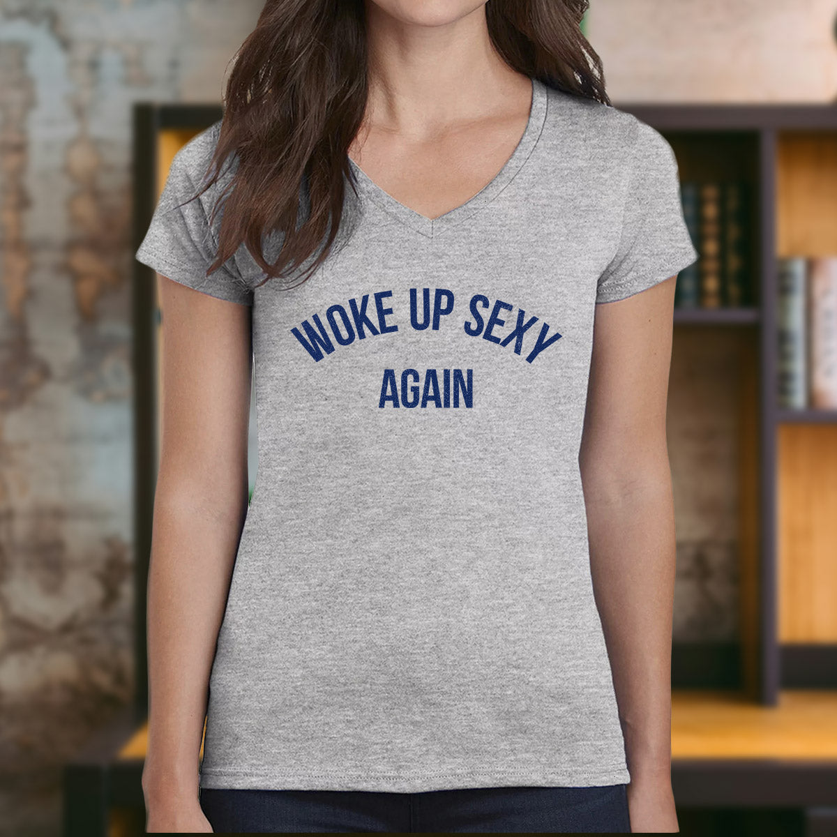 Woman in v-neck grey t-shirt with navy text "Woke Up Sexy Again" standing indoors near bookshelf.