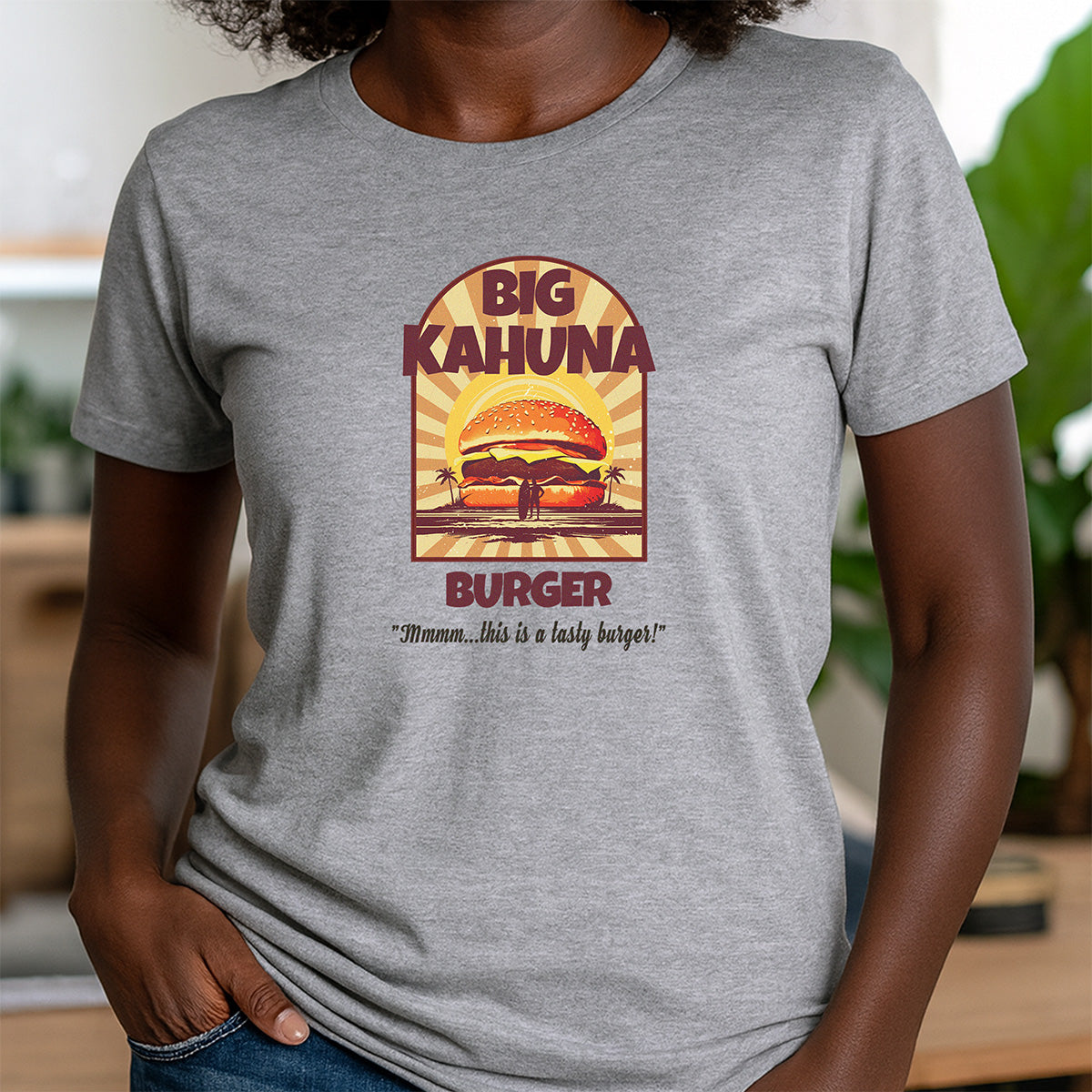 Woman wearing grey crewneck t-shirt with retro Big Kahuna Burger graphic and sunset background