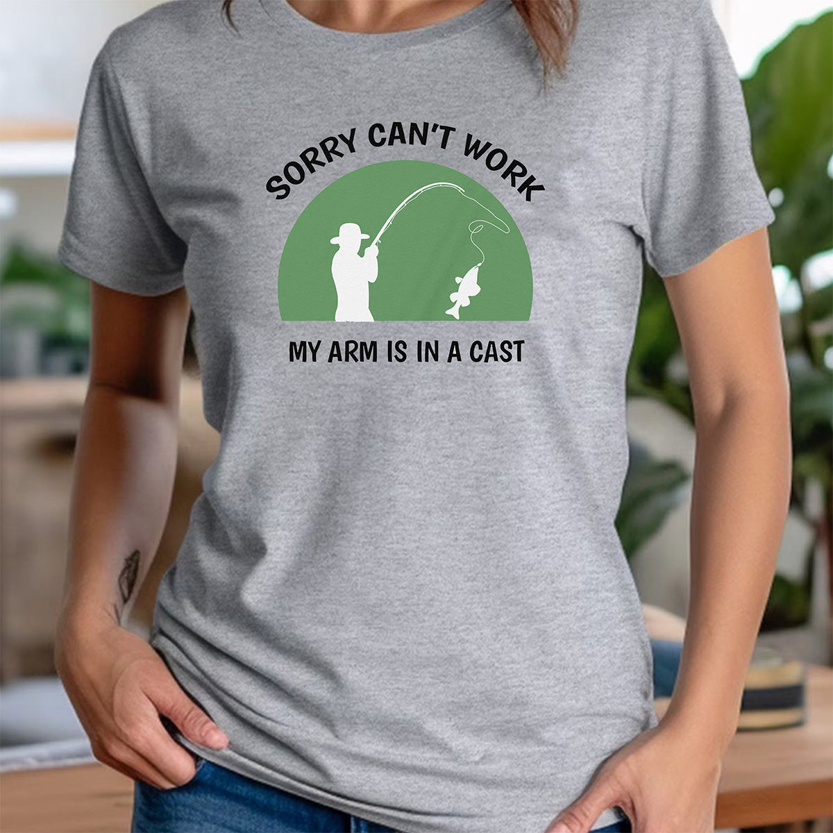 Woman in heather gray crewneck tee with green badge and fisherman silhouette, text Sorry can’t work, my arm is in a cast.