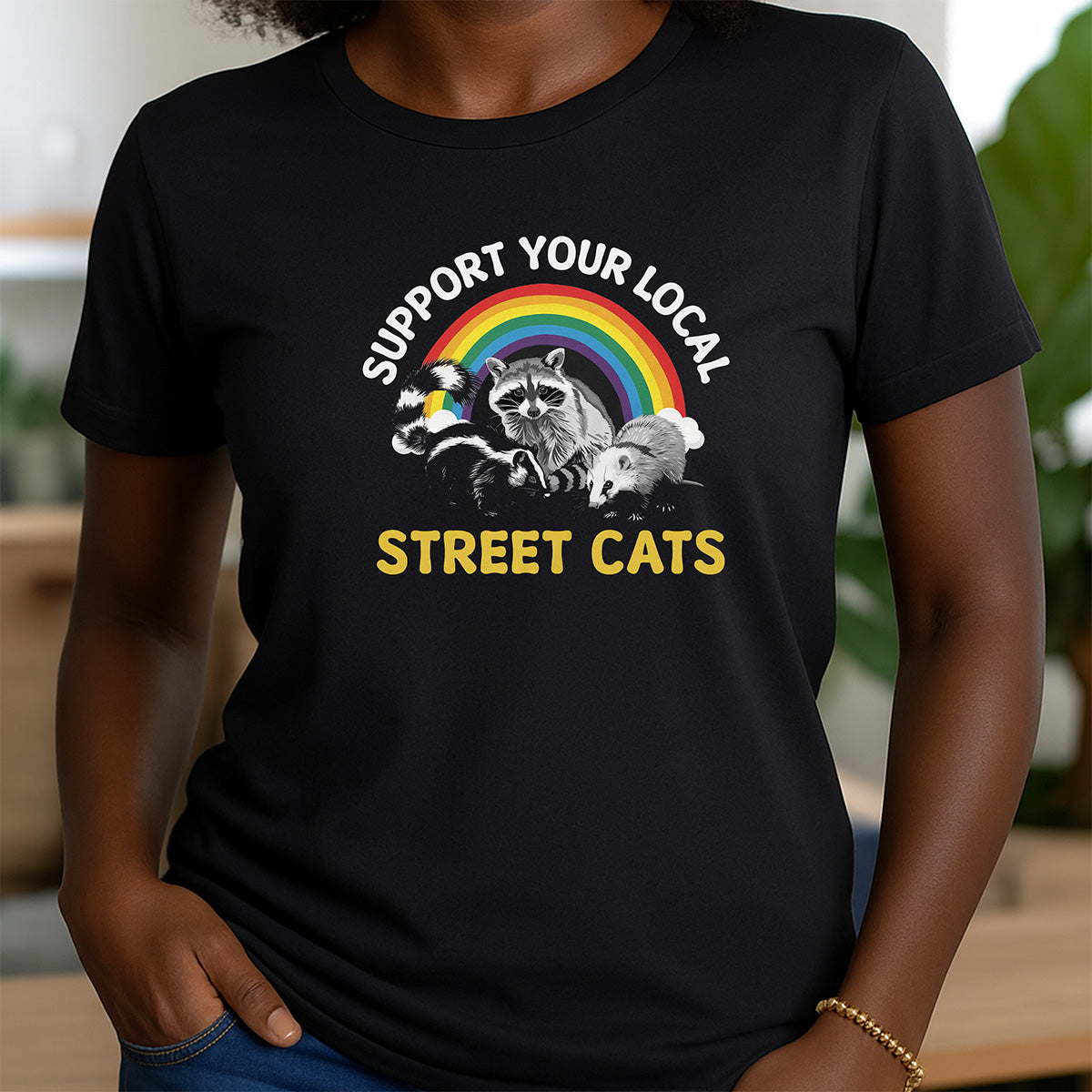 Woman wearing black crewneck shirt with rainbow and raccoon design that reads Support Your Local Street Cats