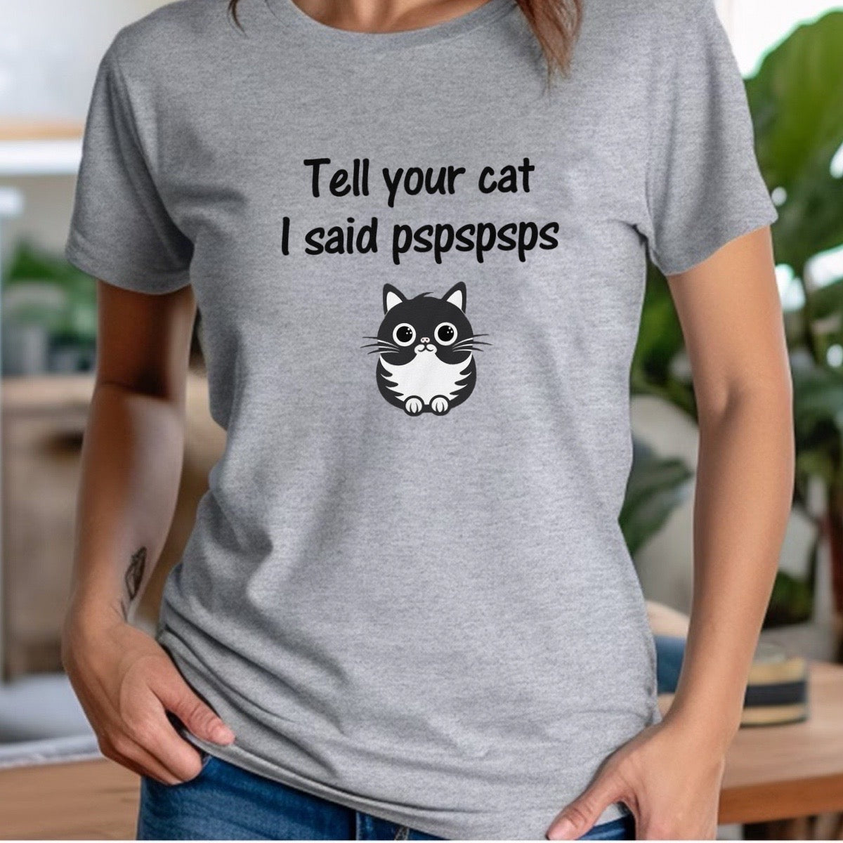 Woman wearing grey cat-themed t-shirt with text “Tell your cat I said pspspsps” and cartoon cat graphic.