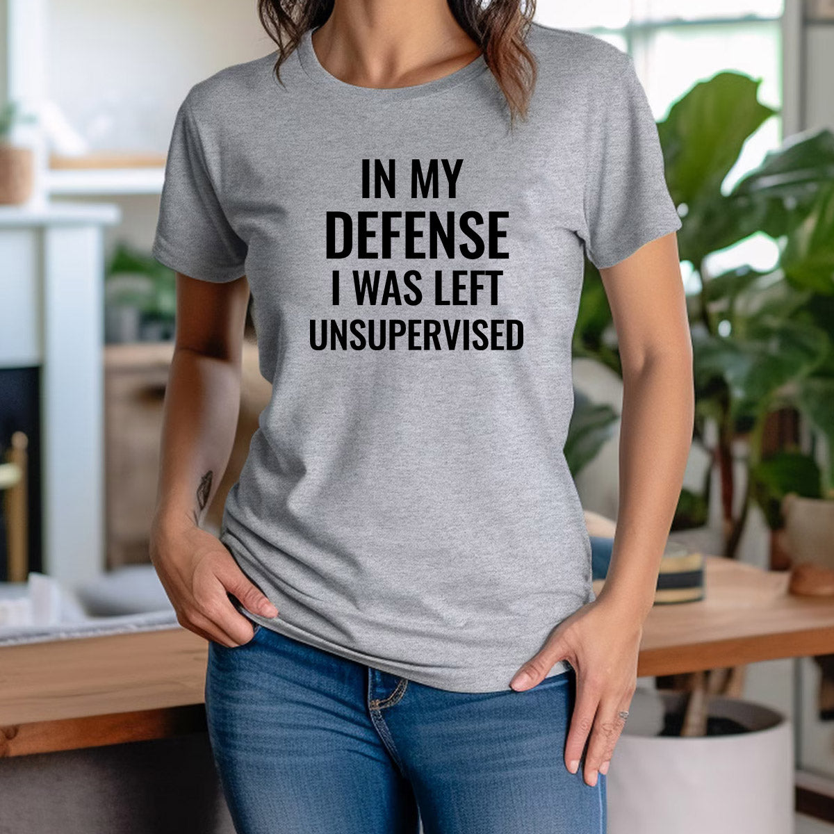 Woman indoors wearing grey graphic t-shirt with bold black quote “In My Defense I Was Left Unsupervised.”