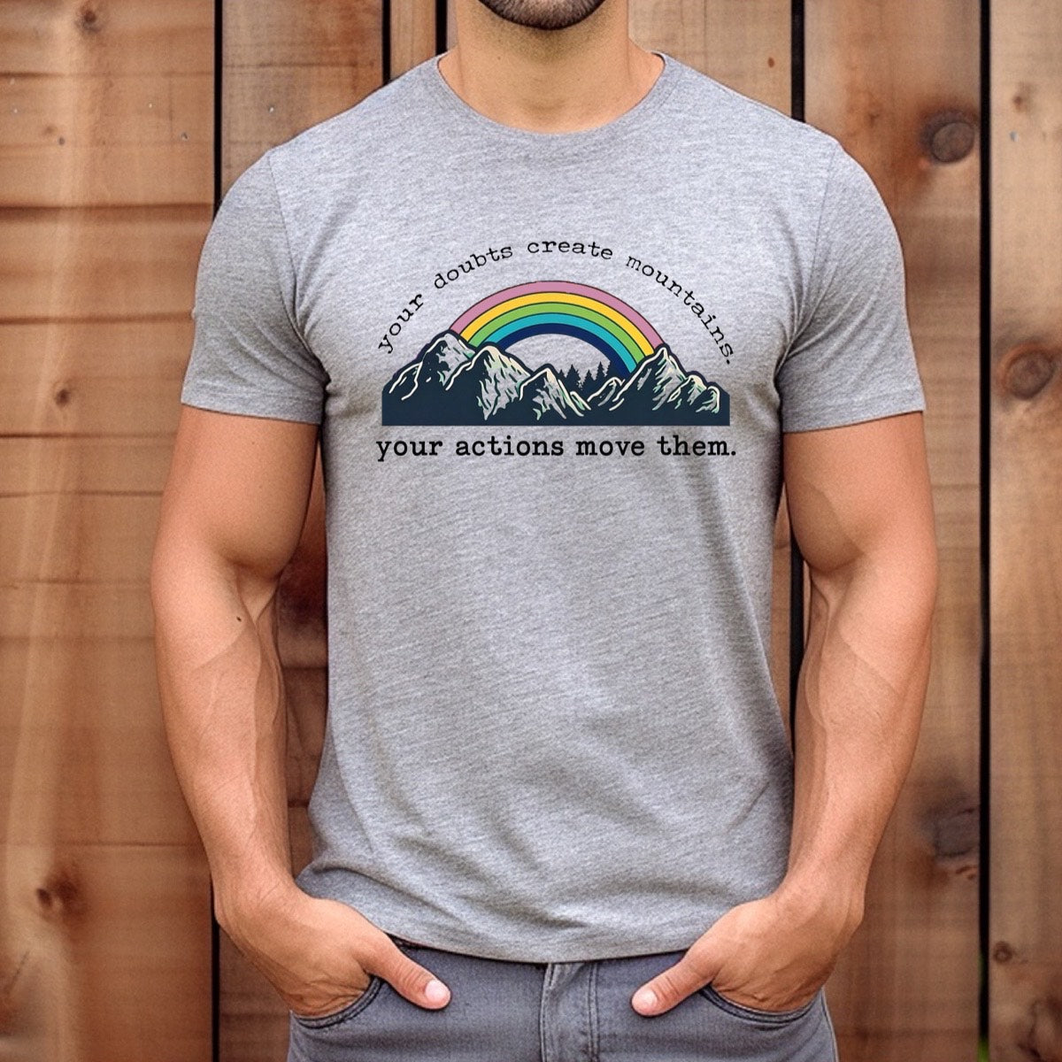 Man wearing heather gray t-shirt with rainbow mountains graphic and quote your doubts create mountains, your actions move them