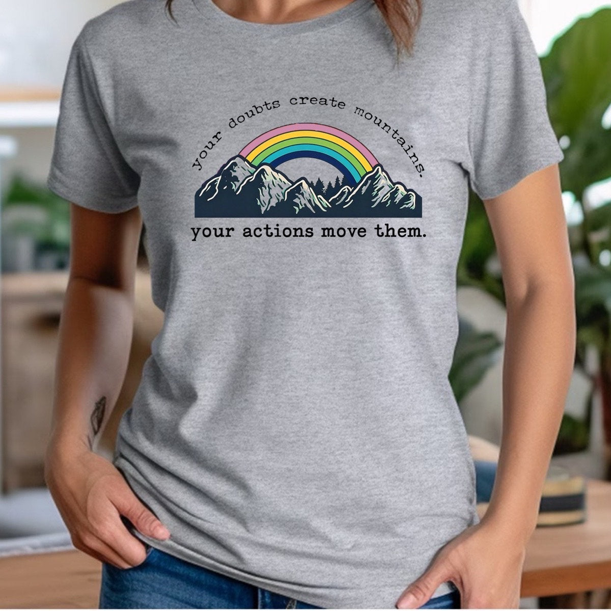 Woman wearing heather gray crewneck t-shirt with rainbow mountains graphic and quote your doubts create mountains, your actions move them
